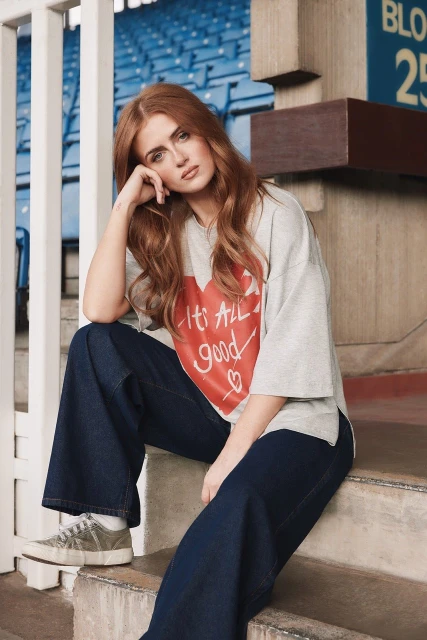 A redhead adult female sits on steps in a gray top with a red heart and blue pants. She is near white railings and a blue stadium backdrop.