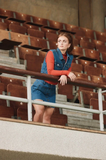 Woman in denim vest and skirt standing in stadium seating area, wearing a red shirt. Natural light, looking away.