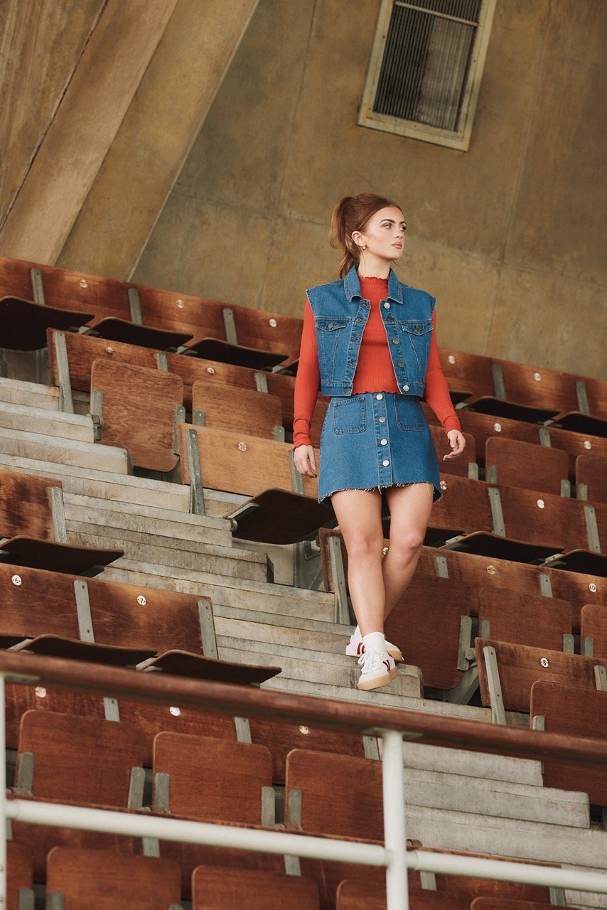 A woman in a denim skirt and vest walks down stairs in an old stadium, looking to her left in a full shot with natural lighting.
