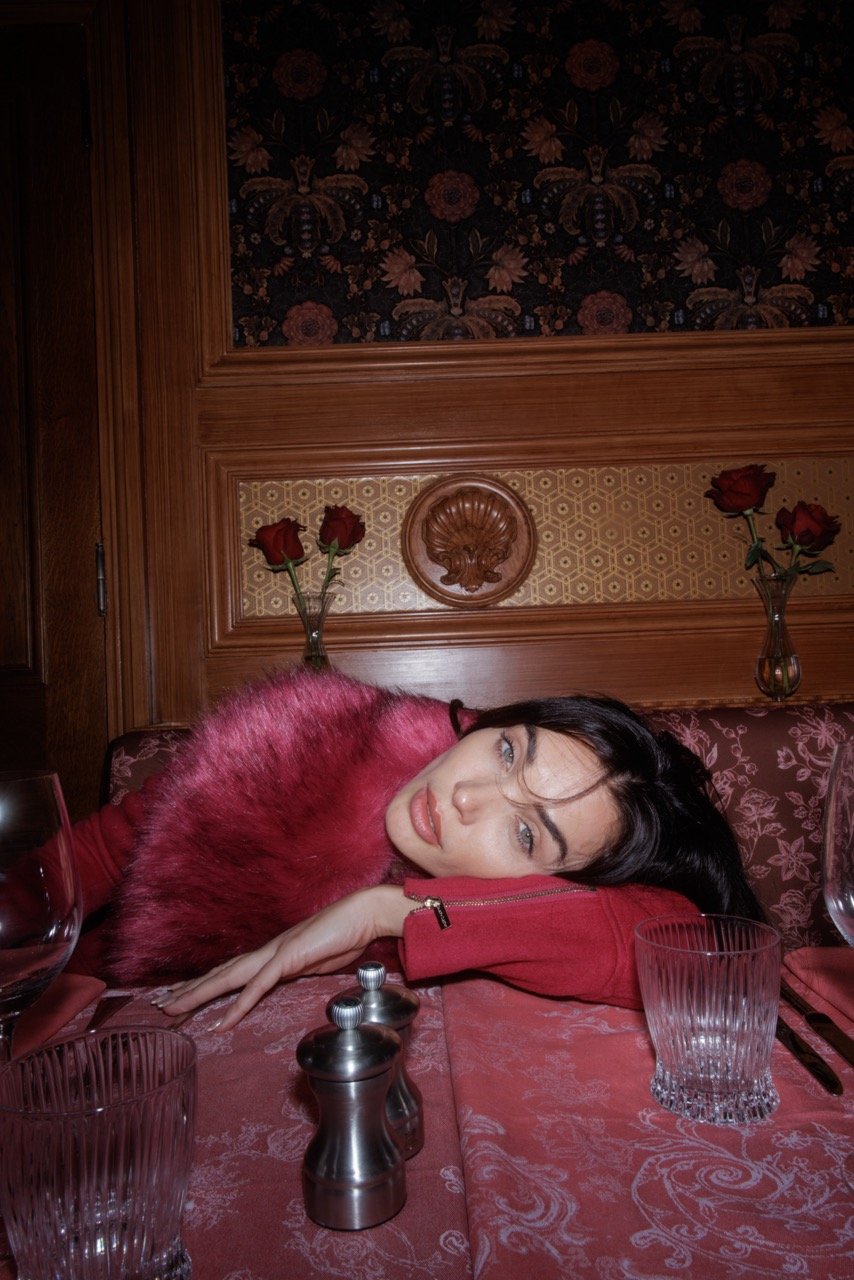 An adult woman with fur shawl poses at a restaurant table with a red tablecloth, glasses, salt shakers, and dark floral wallpaper.