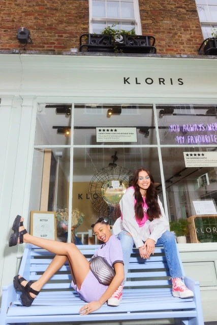 Two young women pose on a light blue bench in front of the Kloris store with large glass windows and a brick facade in an outdoor setting.