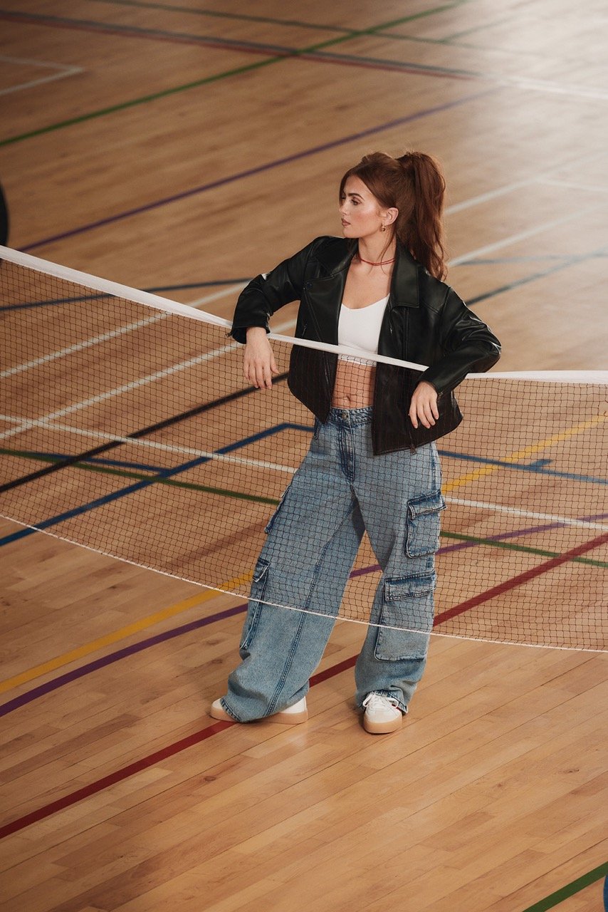 A woman in a black jacket and jeans leans against a badminton net on a wood court with painted lines in a lifestyle fashion shot.