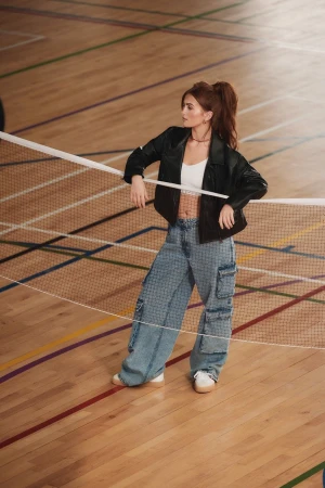 A woman in a black jacket and jeans leans against a badminton net on a wood court with painted lines in a lifestyle fashion shot.