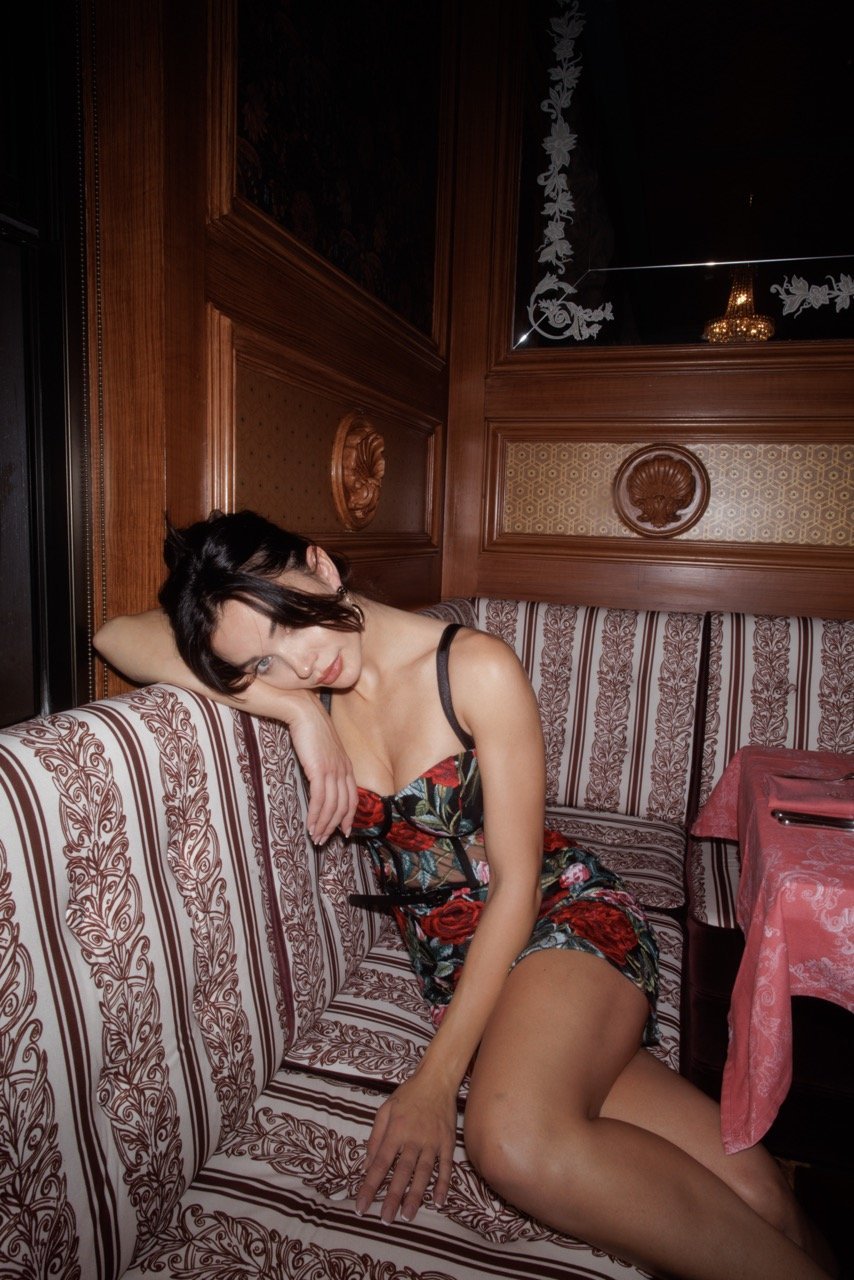 Woman in floral dress reclines on patterned banquette in warm-toned, wood-paneled interior with a table set for dining.