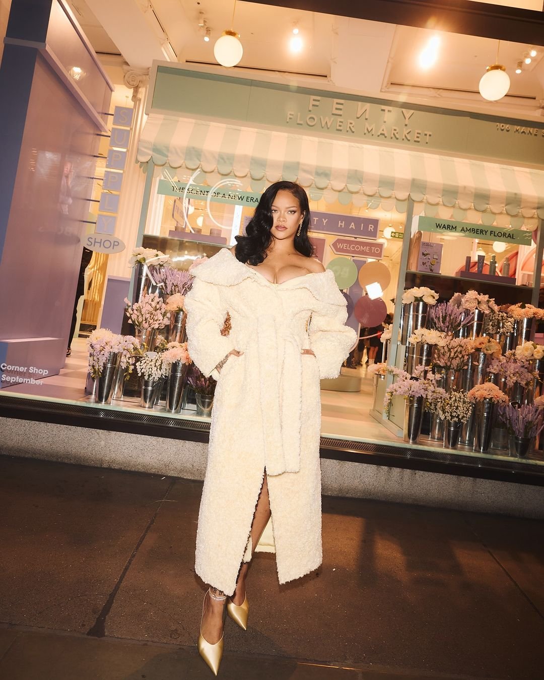 Singer Rihanna poses in front of Fenty Flower Market in a long, off-white, fuzzy coat, gold heels, with wavy hair and makeup.