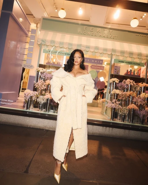 Singer Rihanna poses in front of Fenty Flower Market in a long, off-white, fuzzy coat, gold heels, with wavy hair and makeup.