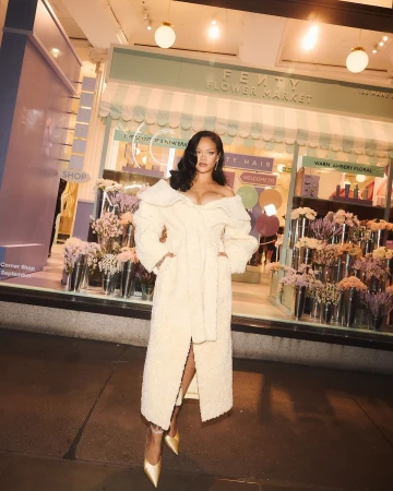 Singer Rihanna poses in front of Fenty Flower Market in a long, off-white, fuzzy coat, gold heels, with wavy hair and makeup.