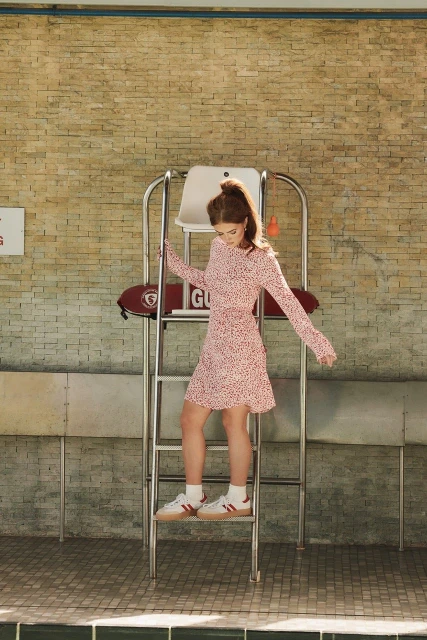 A young adult female in a printed dress stands on a lifeguard tower in a sunlit scene, captured at eye level. Fashion and lifestyle imagery.