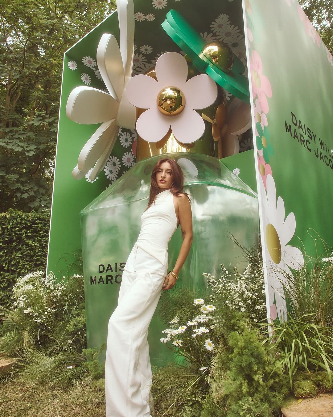 Brunette woman in white outfit poses in front of oversized Marc Jacobs Daisy perfume bottle surrounded by white flowers and greenery.