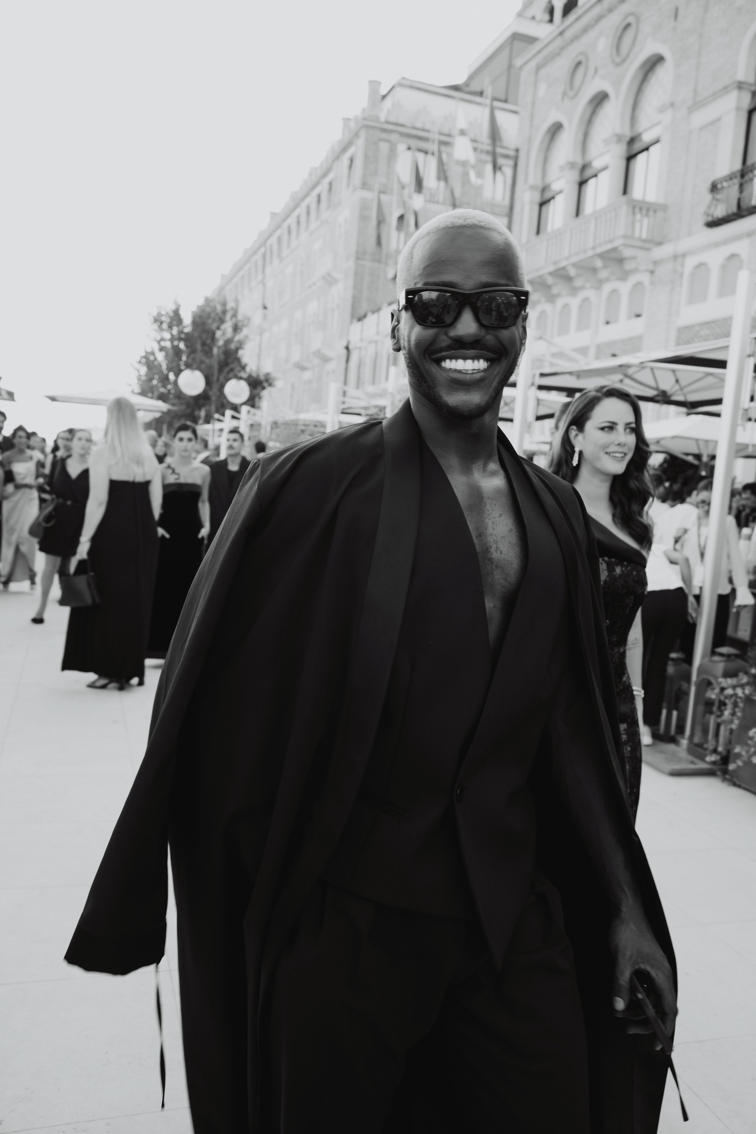 Stylish man in sunglasses smiles while walking in an all-black outfit. He's surrounded by a crowd and classic architecture in the background.