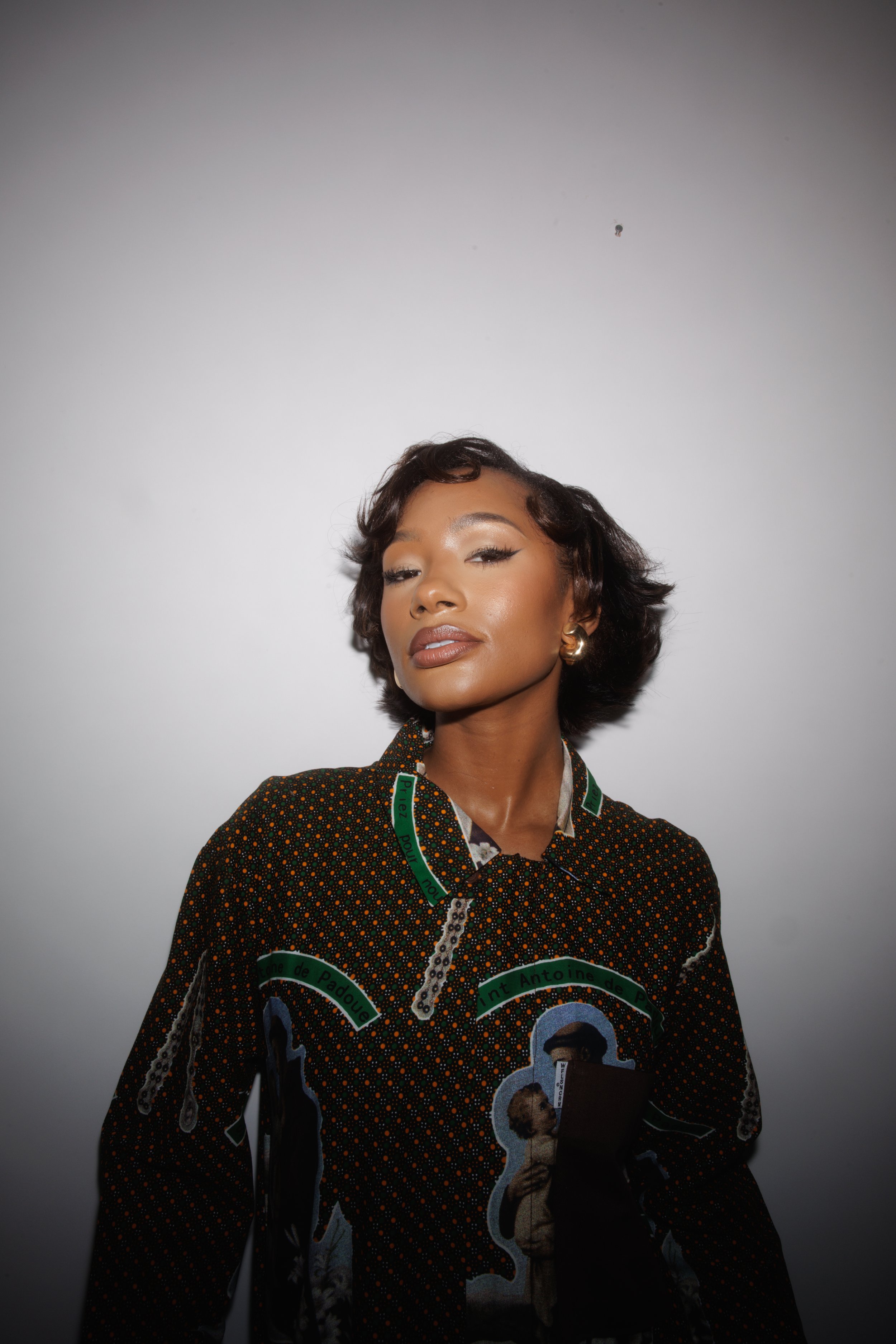 Portrait of a young adult woman with short, dark, wavy hair, wearing a printed long-sleeved top, standing against a plain white wall.