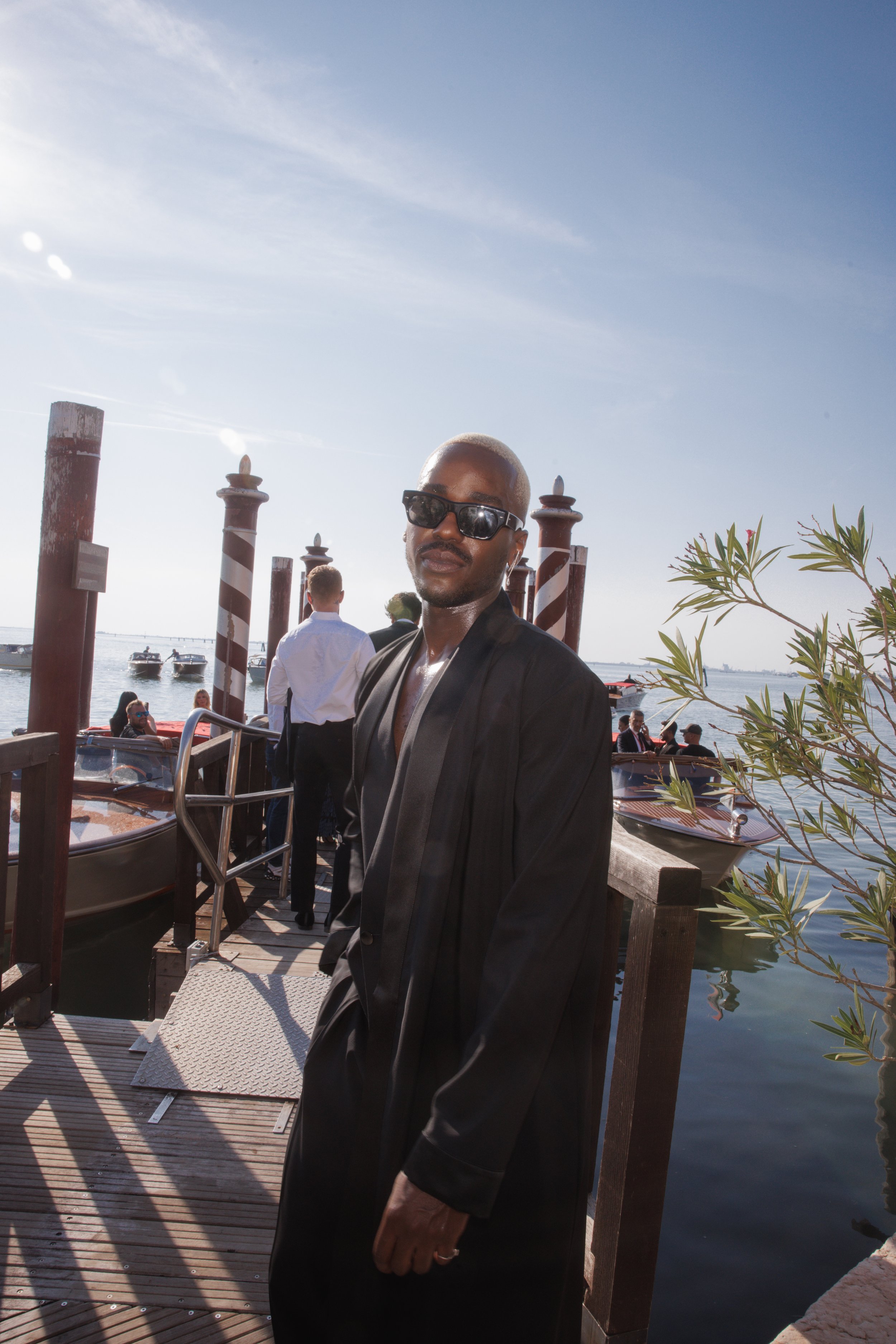 Stylish black man in a long coat and sunglasses stands on a wooden pier near water with boats under a bright sky in Venice, Italy.