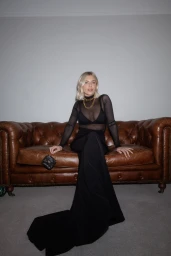 Blonde woman seated on a brown leather tufted sofa wearing a sheer black top and black trousers
