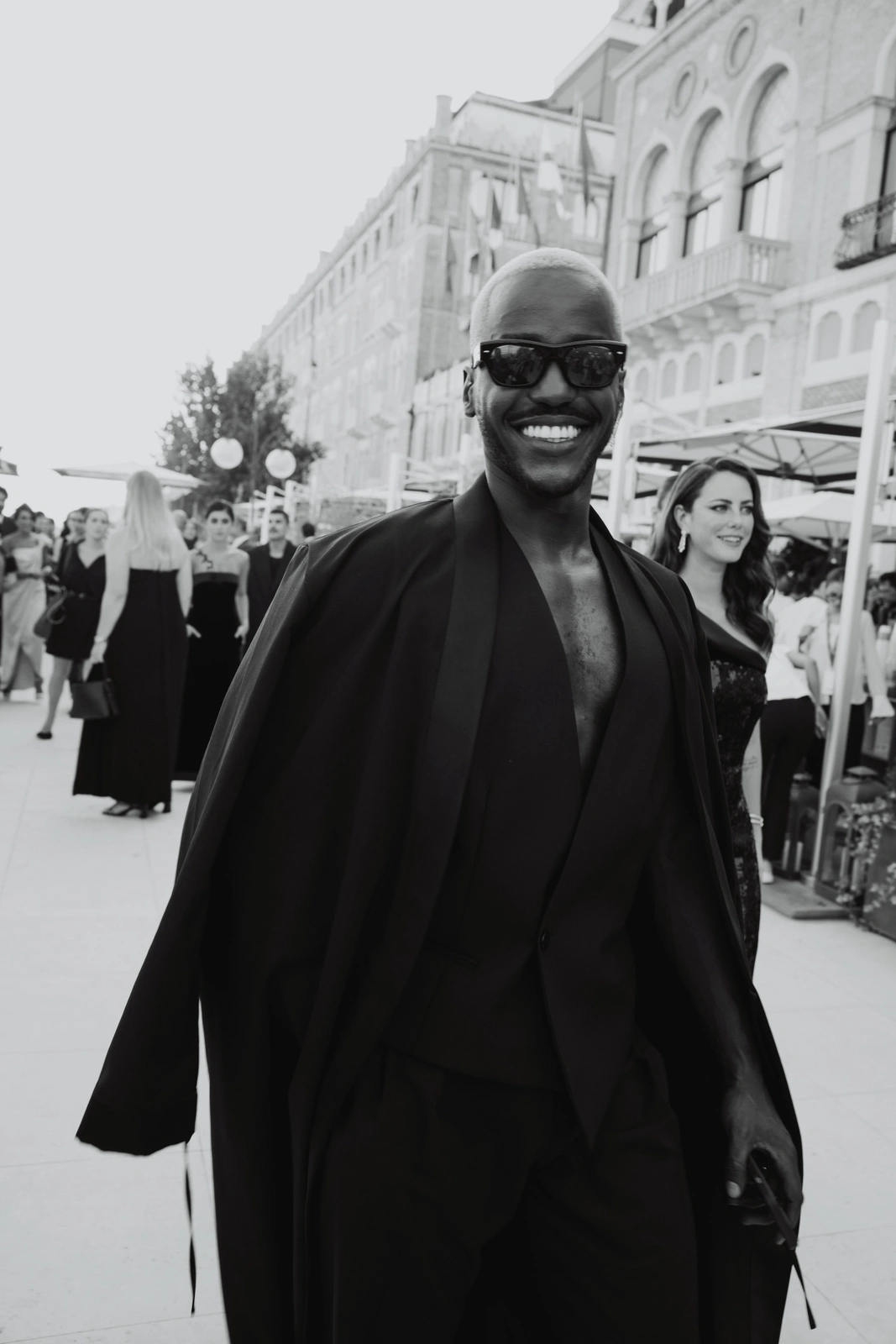 Monochrome portrait of a smiling Black man in dark sunglasses and a black suit jacket draped over his shoulders.