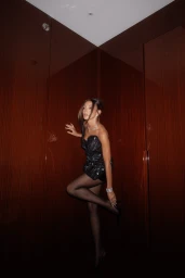 Woman in a black strapless mini dress and sheer tights posing inside a polished wood-paneled elevator.