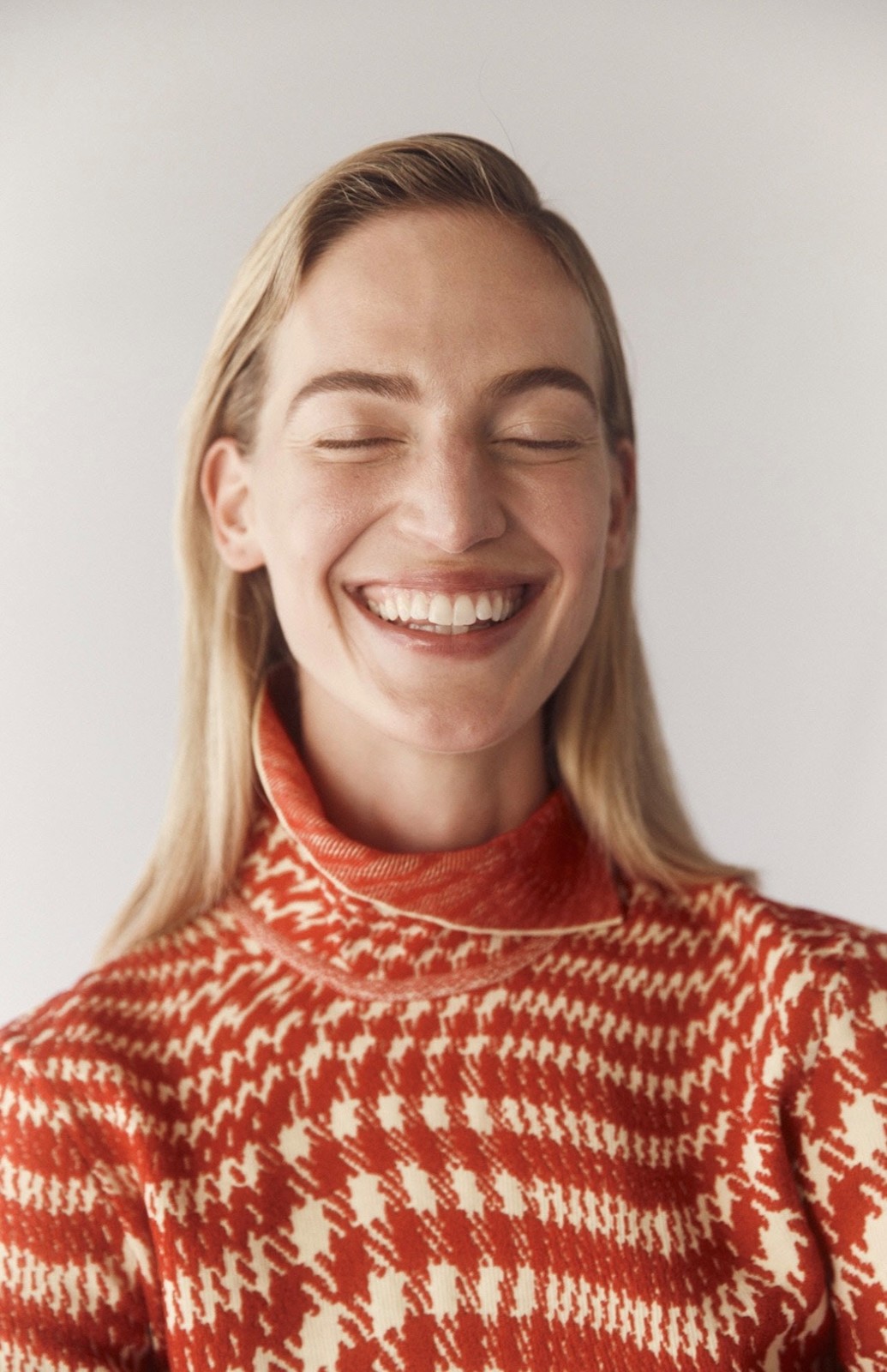 Close-up portrait of a smiling young woman with fair skin and blonde hair, wearing a patterned turtleneck sweater against a white background.
