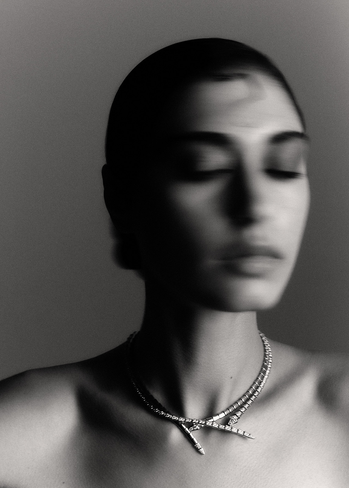 Monochrome portrait of woman with eyes closed, wearing diamond necklace in front of plain studio backdrop.
