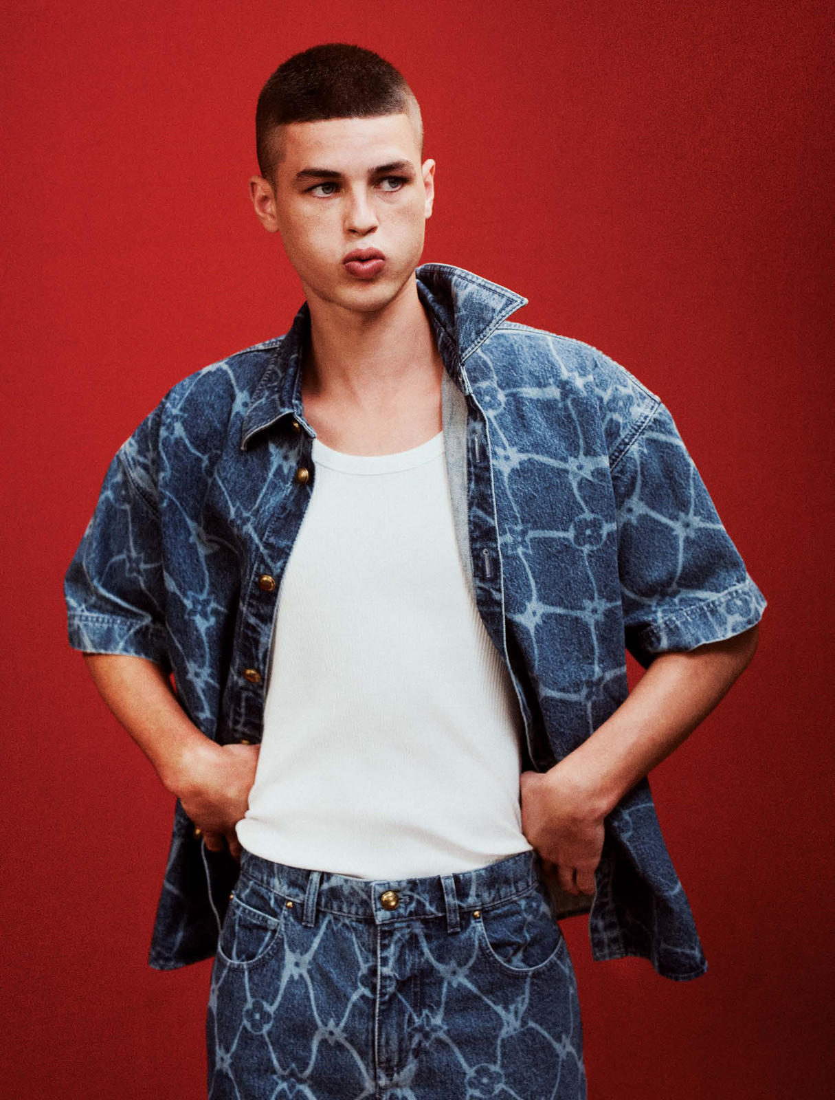Male model poses against a red backdrop wearing a printed denim shirt and jeans with a white tank top, hands casually in pockets.