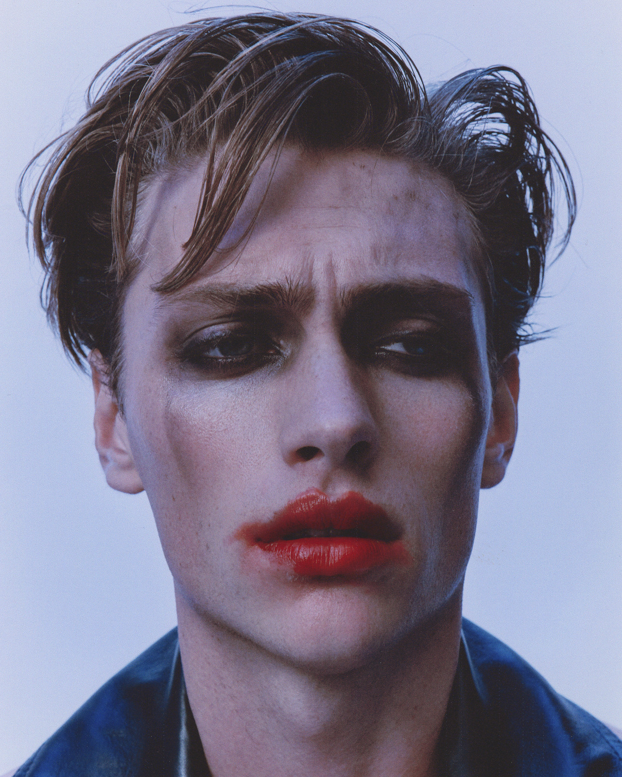 Headshot of a young adult male with smudged makeup around his eyes and red lipstick smeared on his lips, posing against a light backdrop.