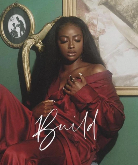 A dark-skinned woman in a burgundy satin outfit is seated in a gold chair against a green wall with paintings in the background.
