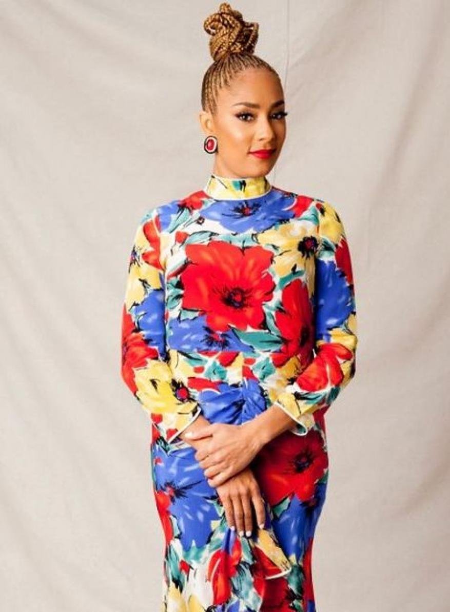 A woman with braided hair stands indoors wearing a vibrant floral dress with red, yellow, and blue flowers and a neutral background.