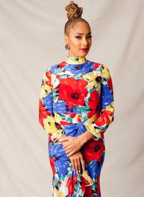 A woman with braided hair stands indoors wearing a vibrant floral dress with red, yellow, and blue flowers and a neutral background.