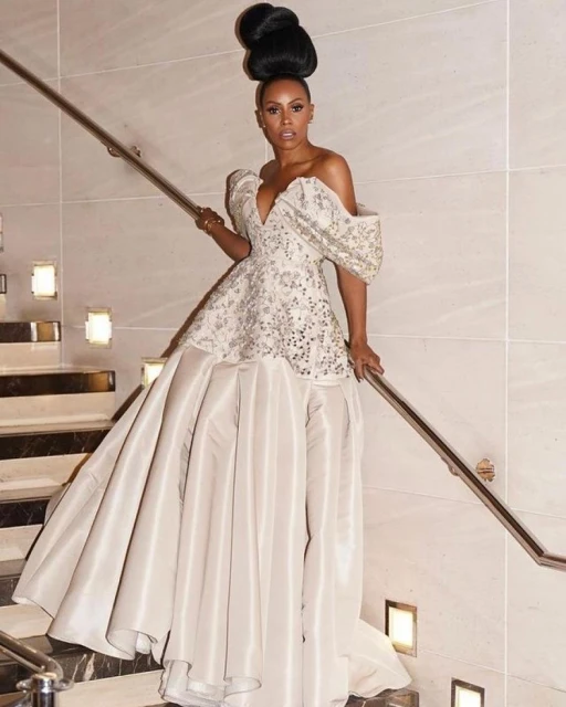 Elegant woman in a formal off-the-shoulder gown poses on stairs with a metal handrail, illuminated by wall-mounted lights.