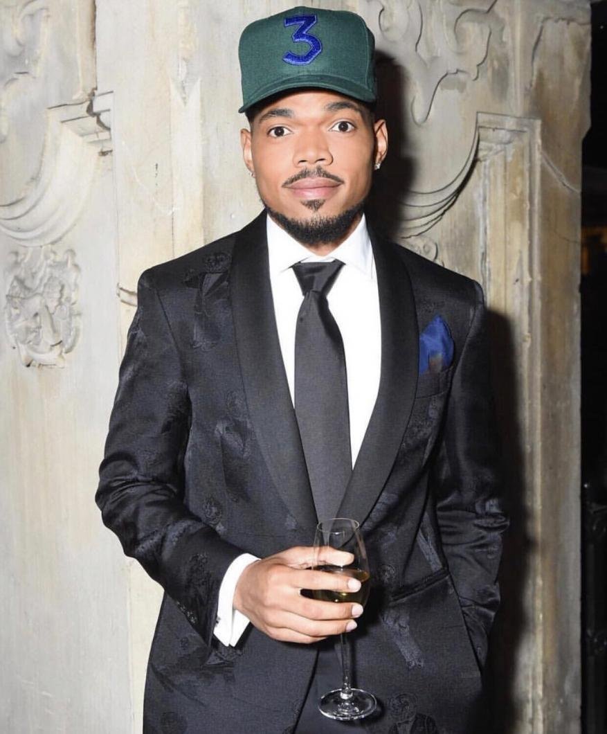 Portrait of Chance the Rapper wearing a green baseball cap and tuxedo, holding a champagne glass at an indoor event.
