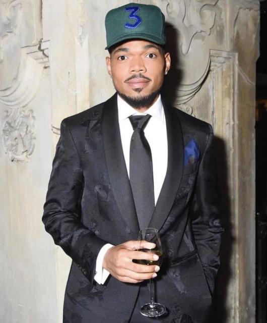 Portrait of Chance the Rapper wearing a green baseball cap and tuxedo, holding a champagne glass at an indoor event.