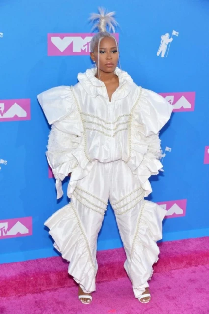 An adult female poses on a pink carpet at the MTV Video Music Awards with a light-blue background wearing a white ruffled outfit.