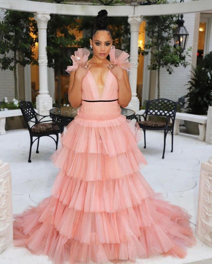 Woman in a peach tulle gown with ruffled shoulders posing in a courtyard with white columns, black iron chairs, and green foliage.