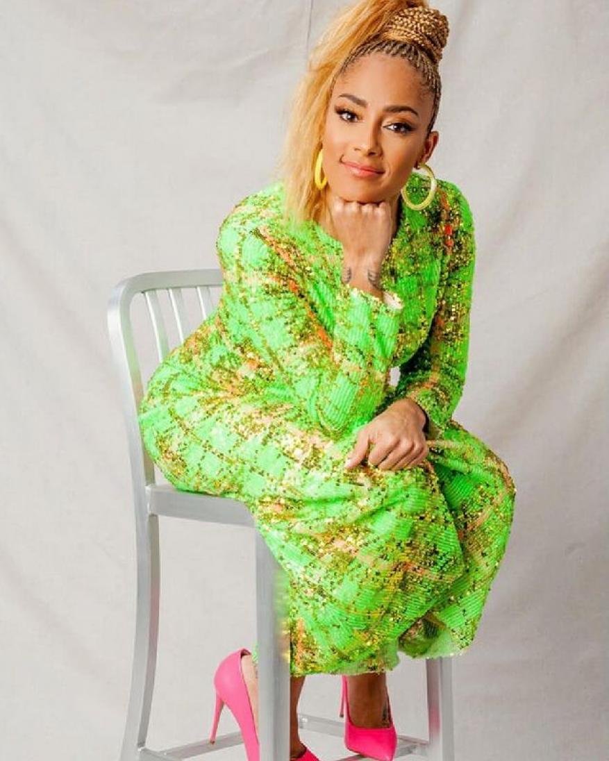 Attractive adult female with a blonde ponytail wearing a sparkling green dress and pink high heels posing on a silver chair in studio.