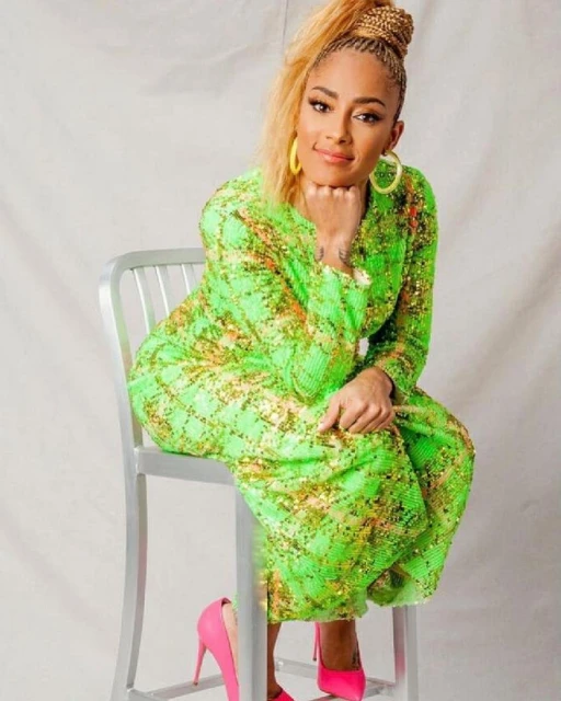 Attractive adult female with a blonde ponytail wearing a sparkling green dress and pink high heels posing on a silver chair in studio.