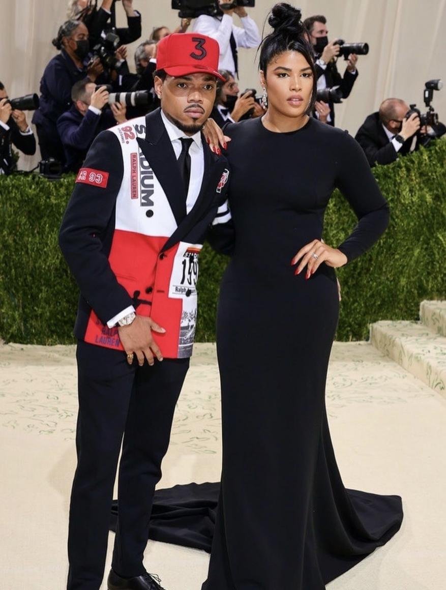 Chance the Rapper and his wife Kirsten Corley pose on a red carpet, surrounded by photographers, wearing formal attire.
