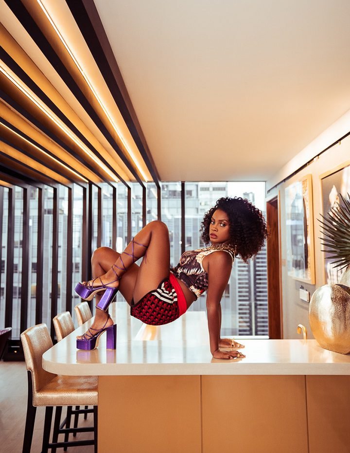 Attractive woman posing on a table in a modern interior setting. She is wearing a patterned top and shorts, and platform heels.