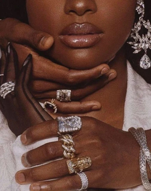 Close-up of dark-skinned person's face and hands adorned with large diamond rings and a diamond earring, held by another's hand.