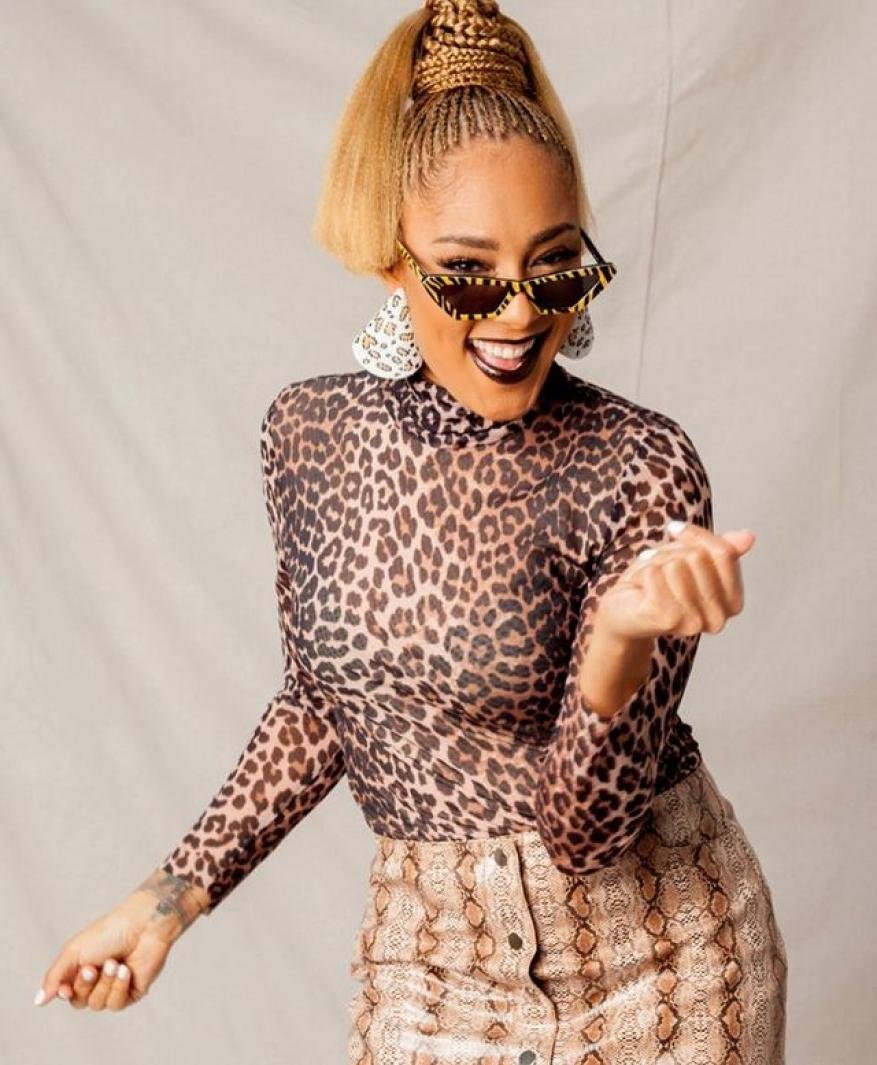 A confident woman with braided blonde hair and sunglasses, wearing a leopard-print top and snakeskin skirt, poses indoors against a neutral backdrop.