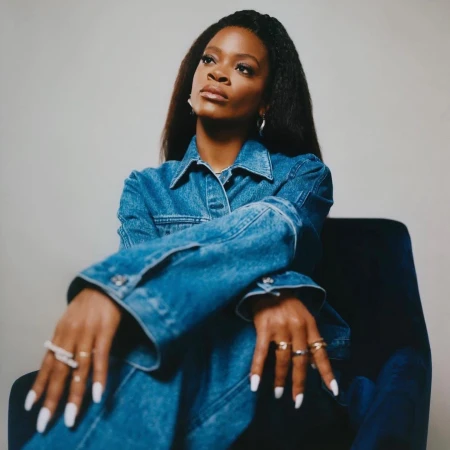 Portrait of a Black woman in a blue denim jacket seated in a dark velvet armchair against a neutral background.