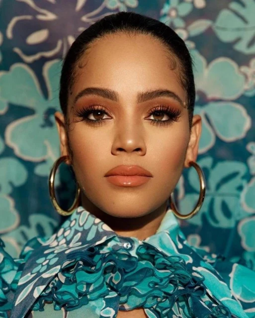Close-up portrait of an adult woman with gold hoop earrings, wearing a turquoise floral top, against a matching floral backdrop.