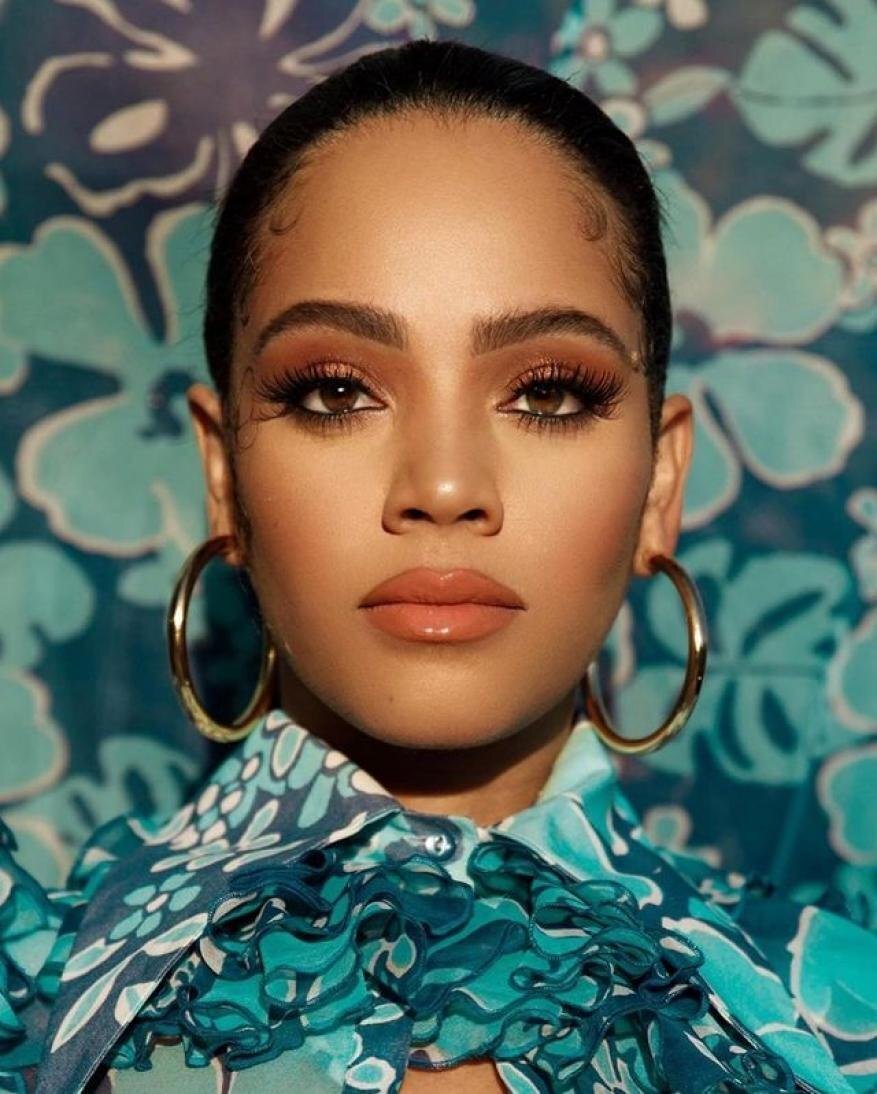 Close-up portrait of a woman with brown hair and eyes, gold hoop earrings, and a floral-patterned blouse against a flower background.