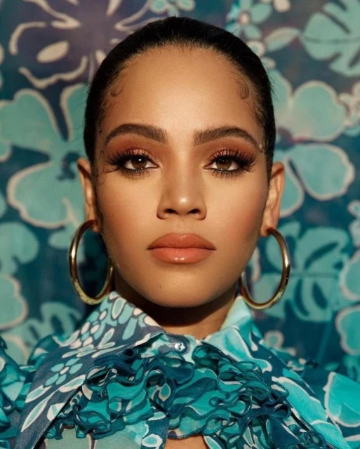 Close-up portrait of a woman with brown hair and eyes, gold hoop earrings, and a floral-patterned blouse against a flower background.