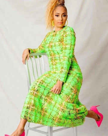 Woman in bright green long-sleeved dress seated on a light grey chair wearing pink high heels against a neutral studio backdrop.