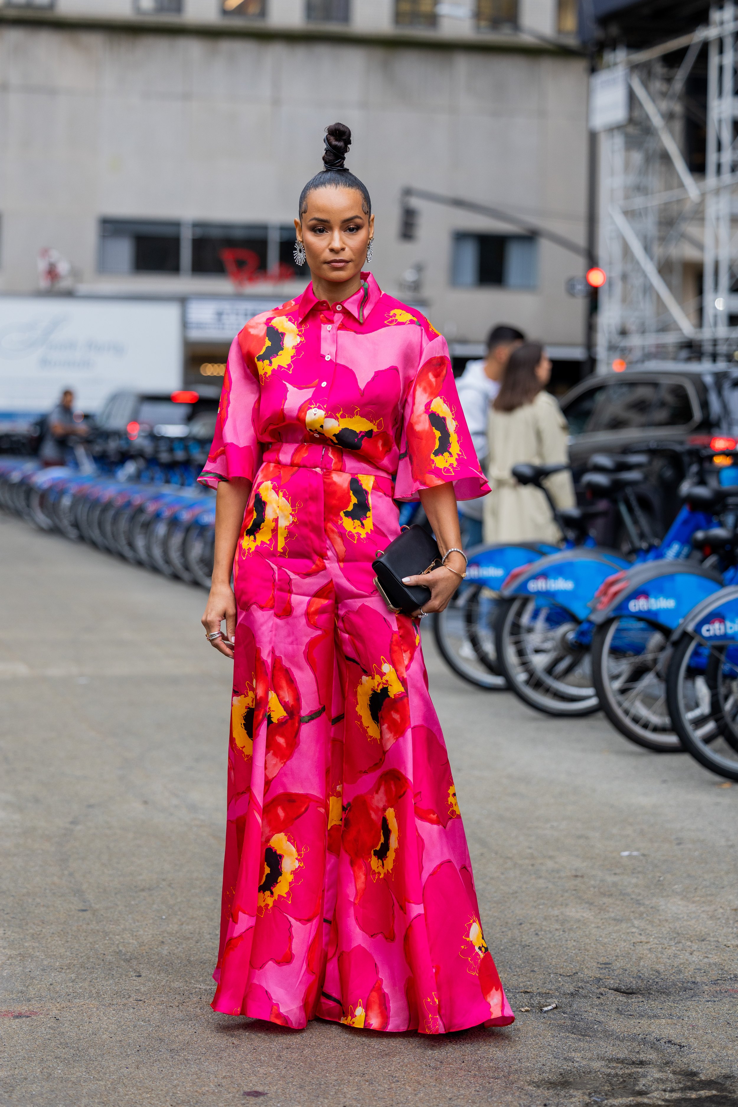 An adult female model stands in an outdoor urban setting, wearing a floral-patterned pink two-piece outfit, holding a black clutch purse.