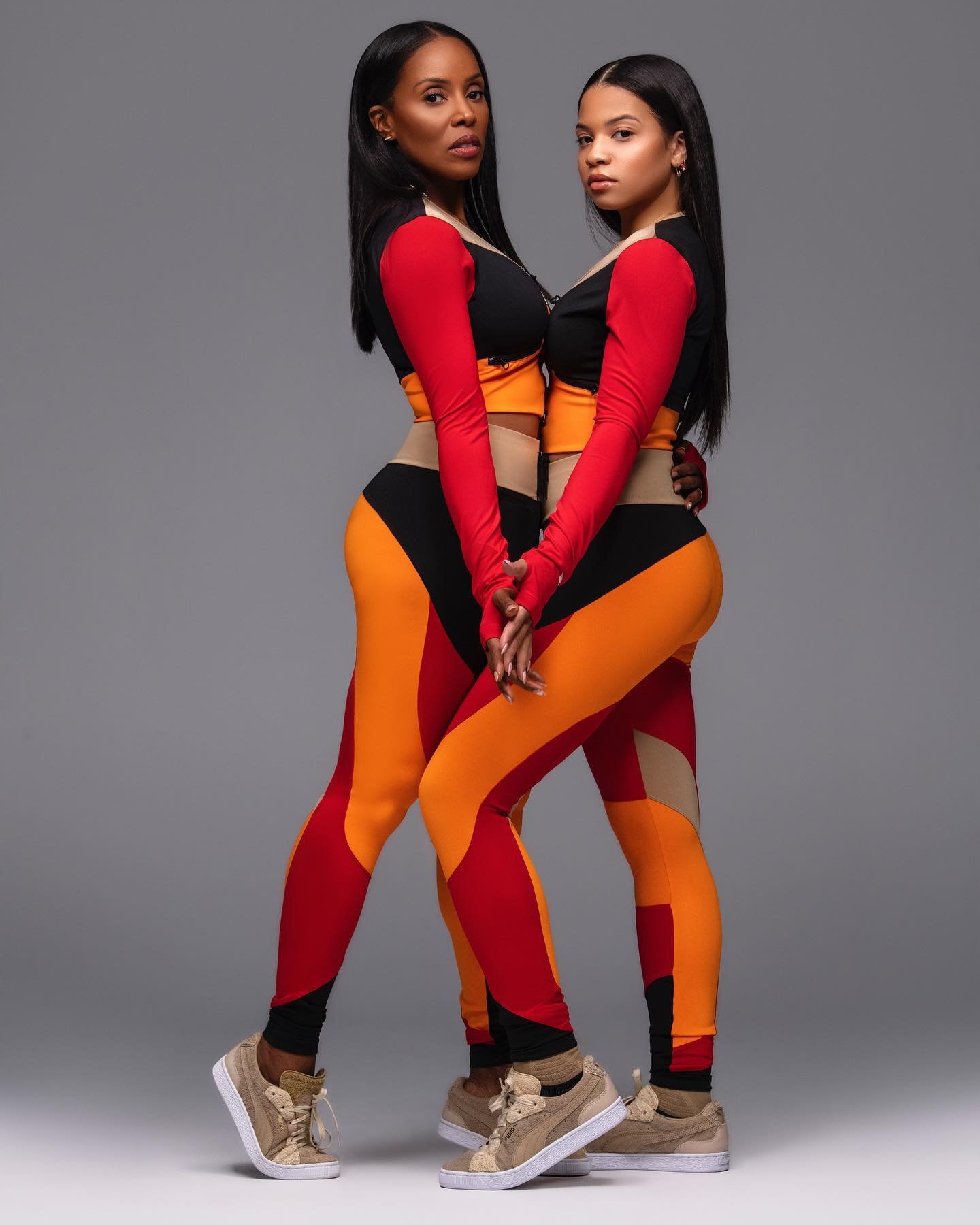 Two women in matching color-blocked athletic wear pose in a studio. The pair have dark hair and tan sneakers, against a plain gray background.