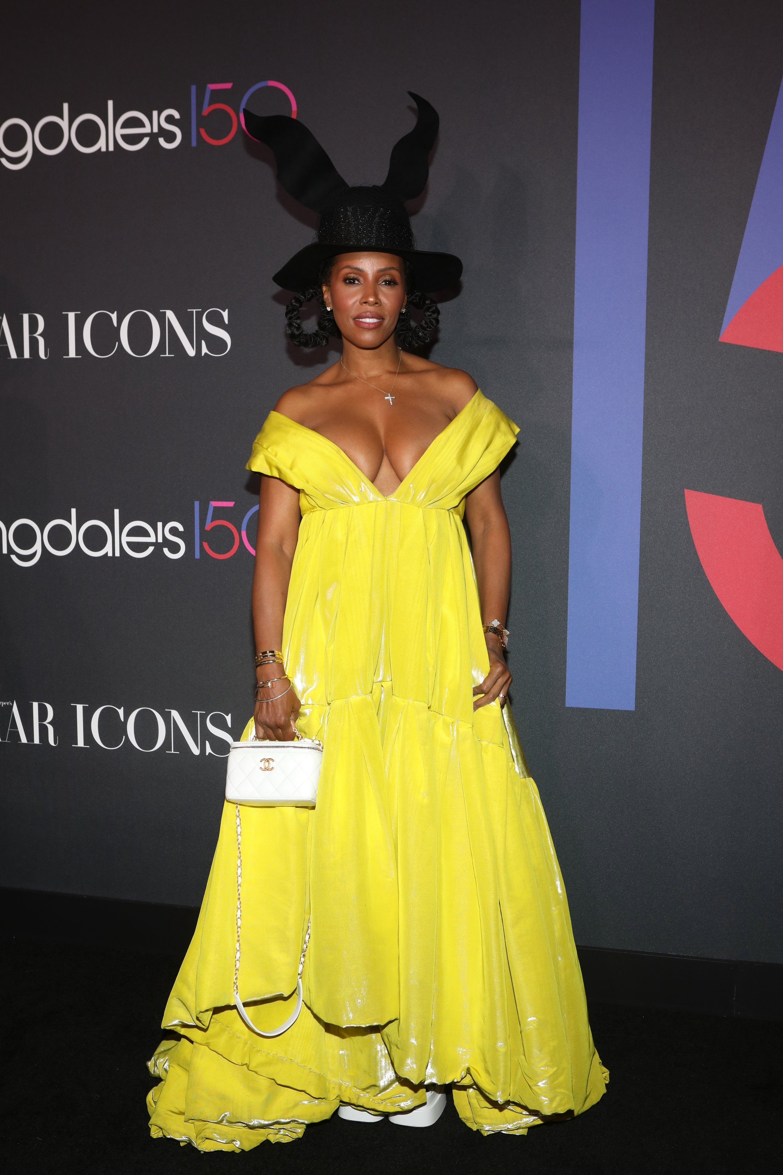 A woman in a yellow dress and a black hat with large rabbit ears poses at an event. She carries a white purse and wears white shoes.
