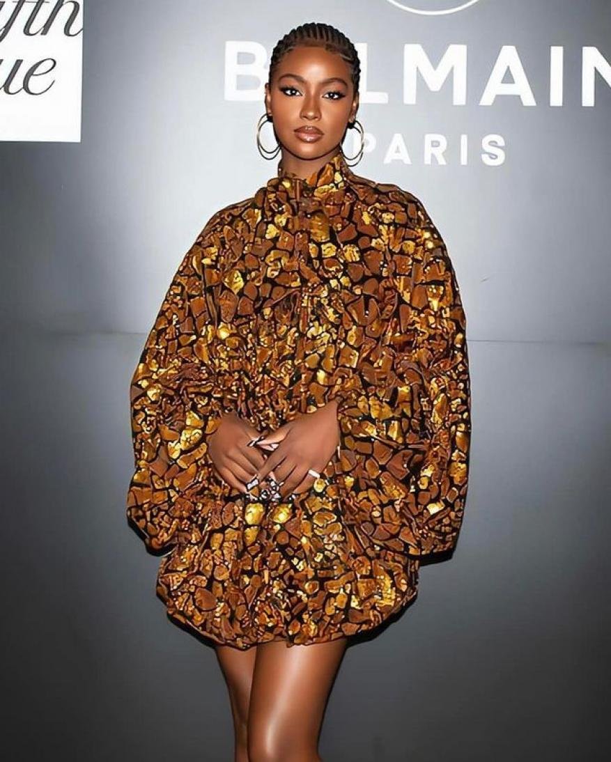 Black woman with braided hair wearing a patterned gold and brown dress standing in front of a Balmain Paris branded wall.