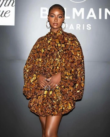 Black woman with braided hair wearing a patterned gold and brown dress standing in front of a Balmain Paris branded wall.