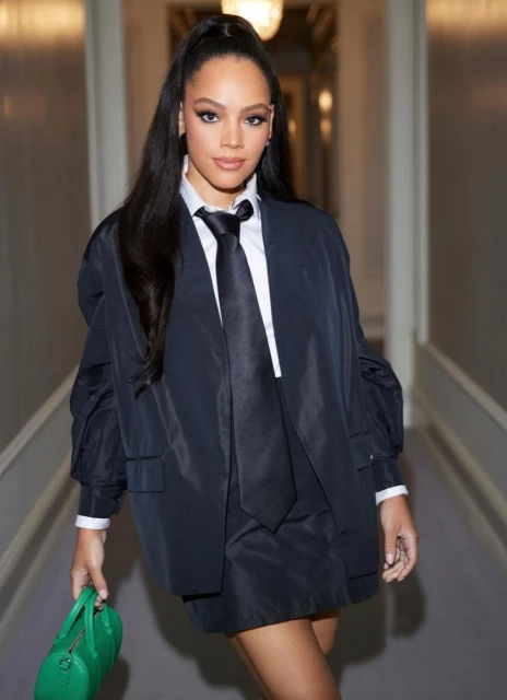 Portrait of a stylish young woman in a black jacket and tie, holding a green handbag, in a brightly lit corridor.