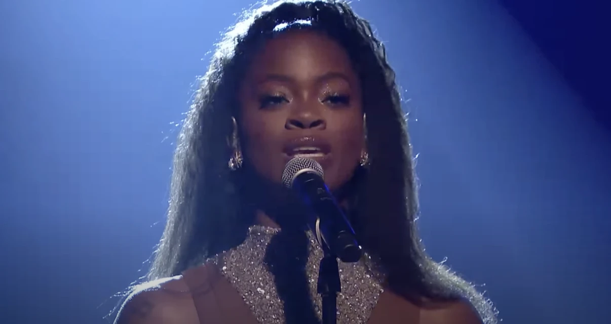 Close-up of a woman singing with a microphone on stage. She has long dark hair, and is wearing a glittery top. Blue backdrop.