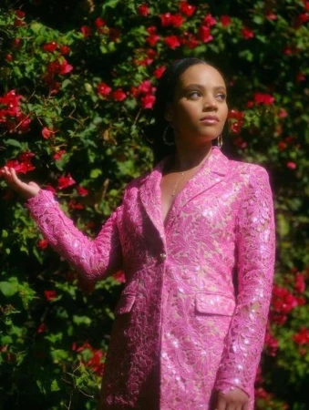 Woman wearing a bright pink lace blazer posing outdoors against a dense green hedge with red flowers in New York